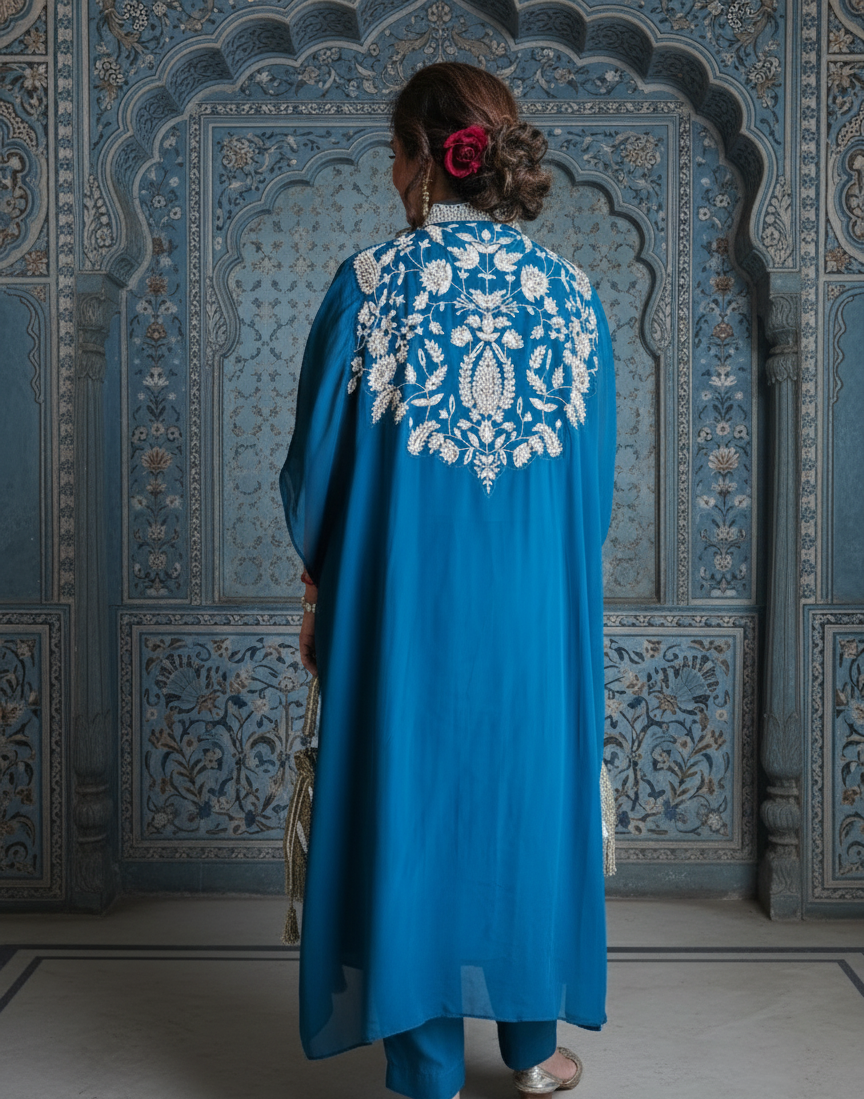 Person wearing a blue embroidered outfit standing in front of an ornate blue wall.